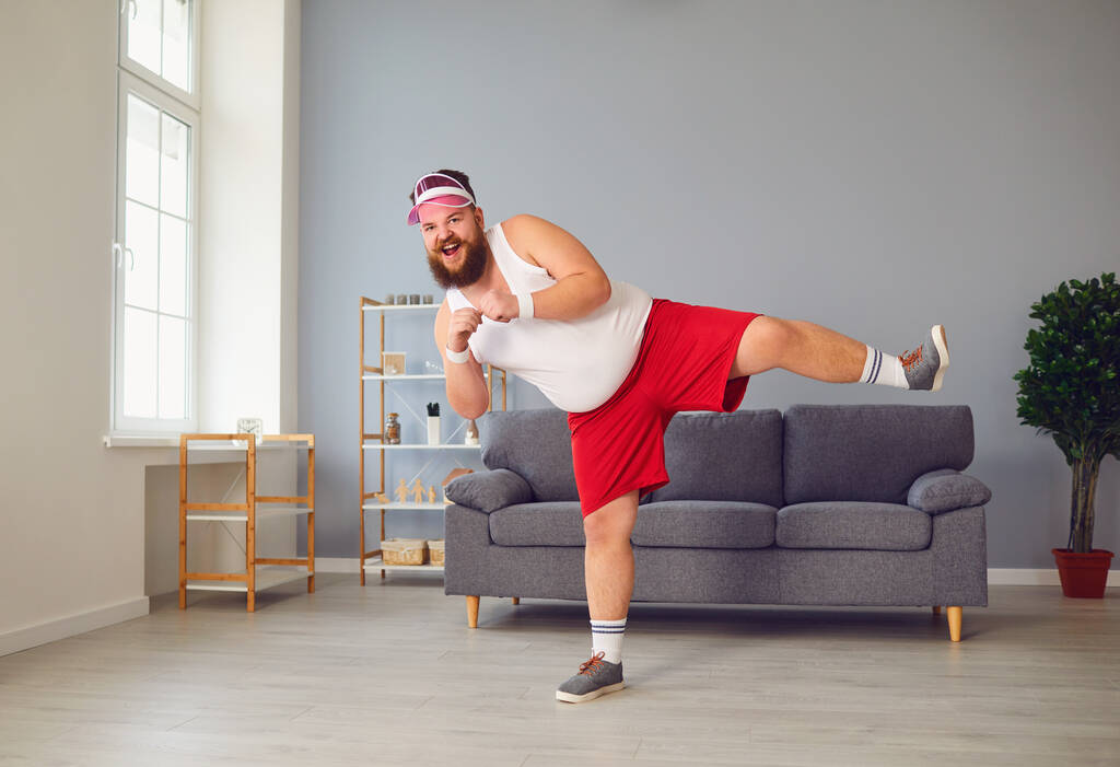 Grappige dikke man in rode broek en wit t-shirt glimlachend oefeningen doen in de kamer - Foto, afbeelding