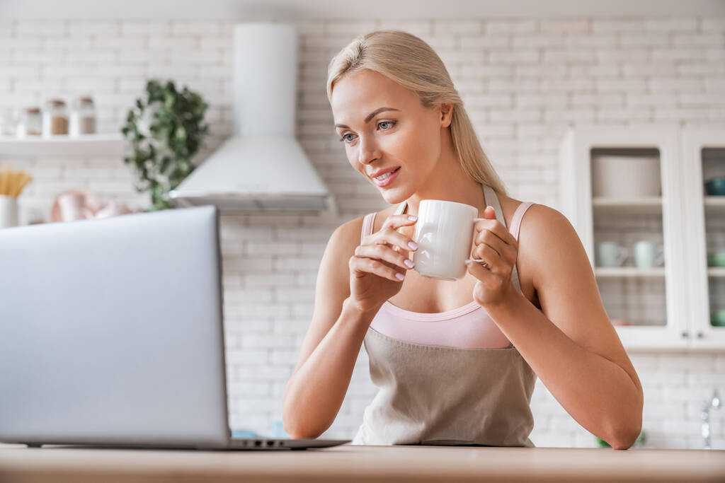 Mulher com xícara de café e laptop na cozinha
 - Foto, Imagem