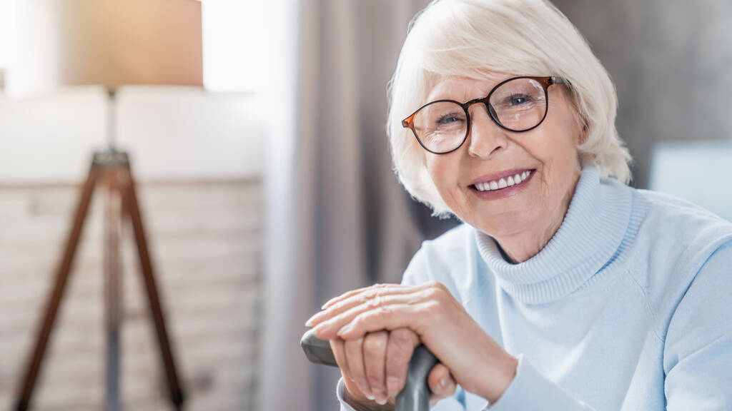 Porträt einer glücklichen reifen Frau mit Brille und Stock, während sie zu Hause auf dem Sofa sitzt - Foto, Bild