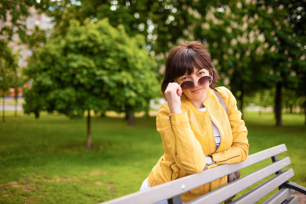 Bella dai capelli rossi attraente donna caucasica in abiti casual e occhiali da sole in posa sulla panchina del parco nella soleggiata giornata primaverile. Concetto di seconda giovinezza e salute durante la menopausa
 - Foto, immagini