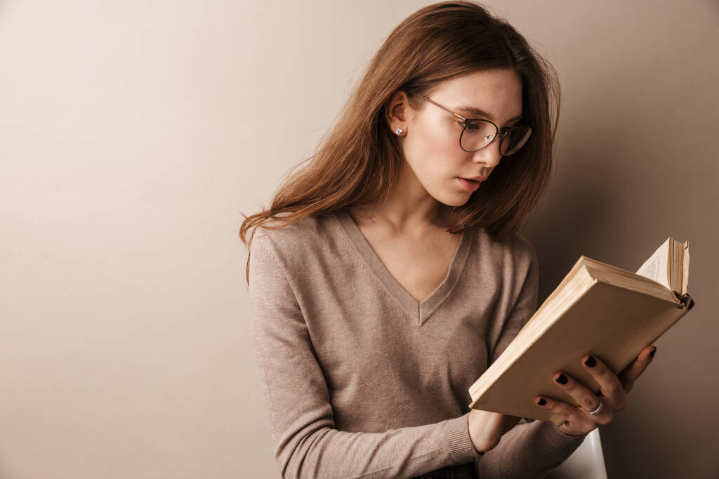 Foto di affascinante donna seria in occhiali libro di lettura e seduta sulla sedia isolata sopra parete grigia
 - Foto, immagini