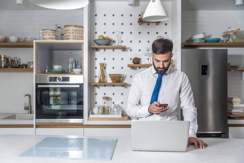 Jeune homme d'affaires barbu en chemise avec cravate au cou debout dans la cuisine tenant le téléphone et lisant la statistique sur l'ordinateur portable attendant le petit déjeuner avant d'aller au travail dans l'entreprise - Photo, image