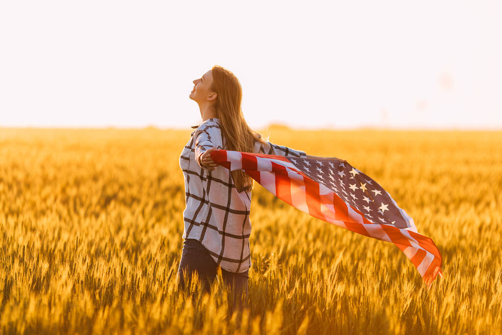 młoda szczęśliwa dziewczyna biegnie i skacze z otwartymi ramionami przez pole pszenicy. Posiadanie flagi USA - Zdjęcie, obraz