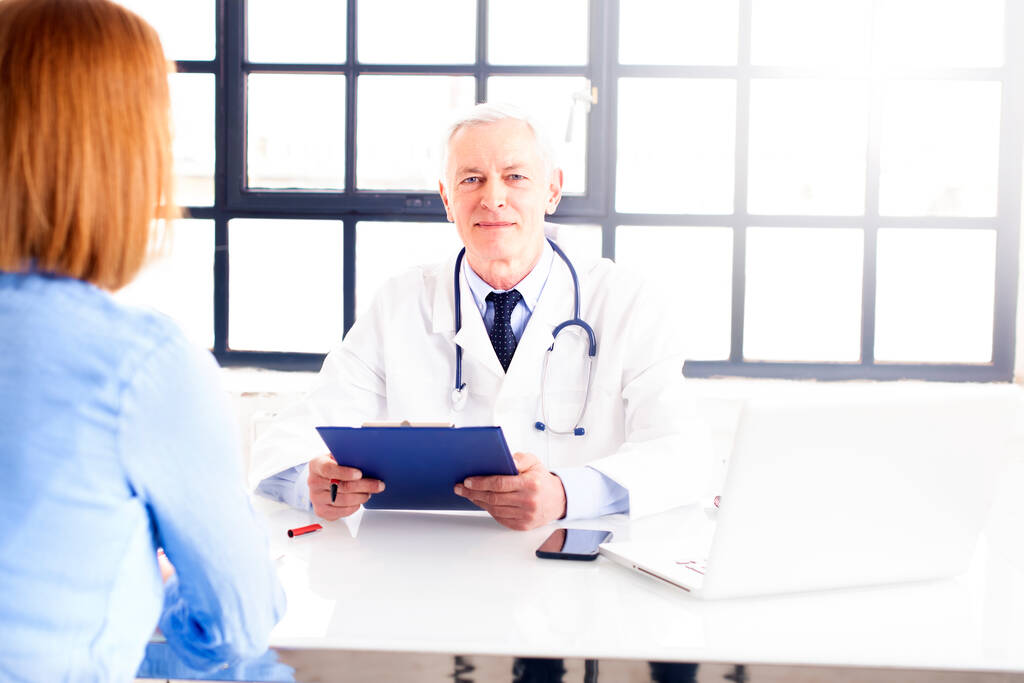 Orta yaşlı erkek doktorun, doktorların odasında masasında otururken hastasıyla konuşması. - Fotoğraf, Görsel