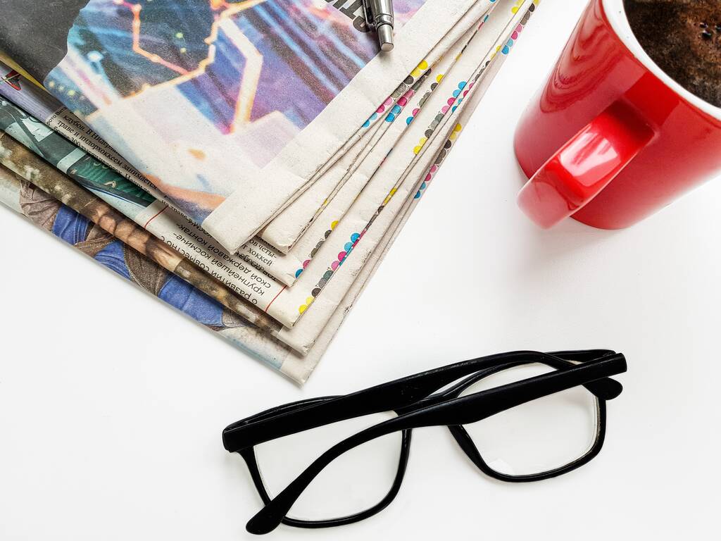 Una pila di vecchi giornali, occhiali neri, una penna e una tazza rossa di caffè nero su sfondo bianco
. - Foto, immagini