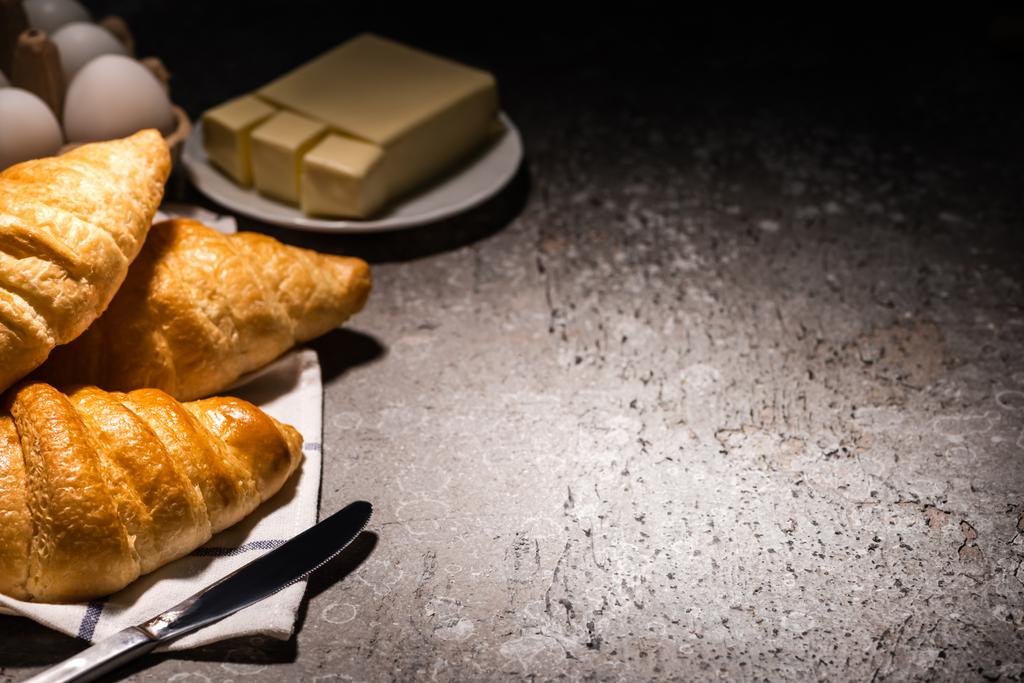 selective focus of fresh baked croissants with knife on towel near butter and eggs on concrete grey surface in dark - Photo, Image