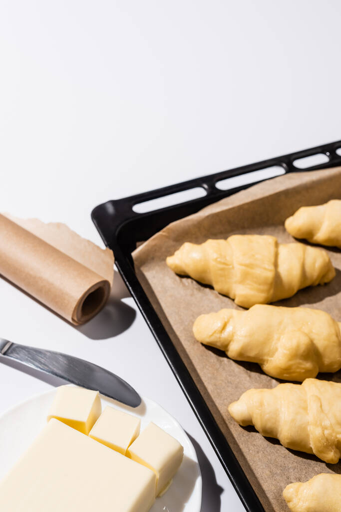 selectieve focus van rauwe croissants op bakplaat bij perkamentpapier, boter, mes op witte achtergrond - Foto, afbeelding