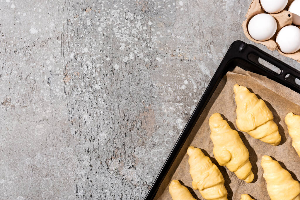 Ansicht von ungekochten Croissants auf Backblech in der Nähe von Eiern auf betongrauer Oberfläche - Foto, Bild
