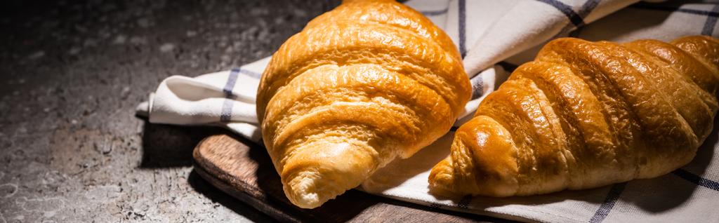 fresh baked croissants on towel and wooden cutting board on concrete grey surface in dark, panoramic shot - Photo, Image