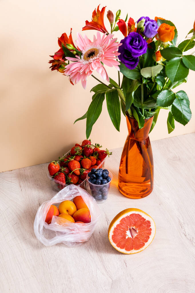 Blumen- und Fruchtkomposition mit Bouquet in Vase, Beeren, Grapefruit und Aprikosen auf Holzoberfläche auf beigem Hintergrund - Foto, Bild