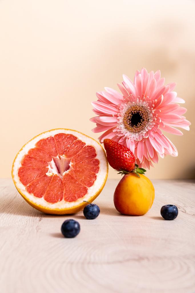 floral and fruit composition with pink gerbera, berries, grapefruit and apricot on wooden surface isolated on beige - Photo, Image