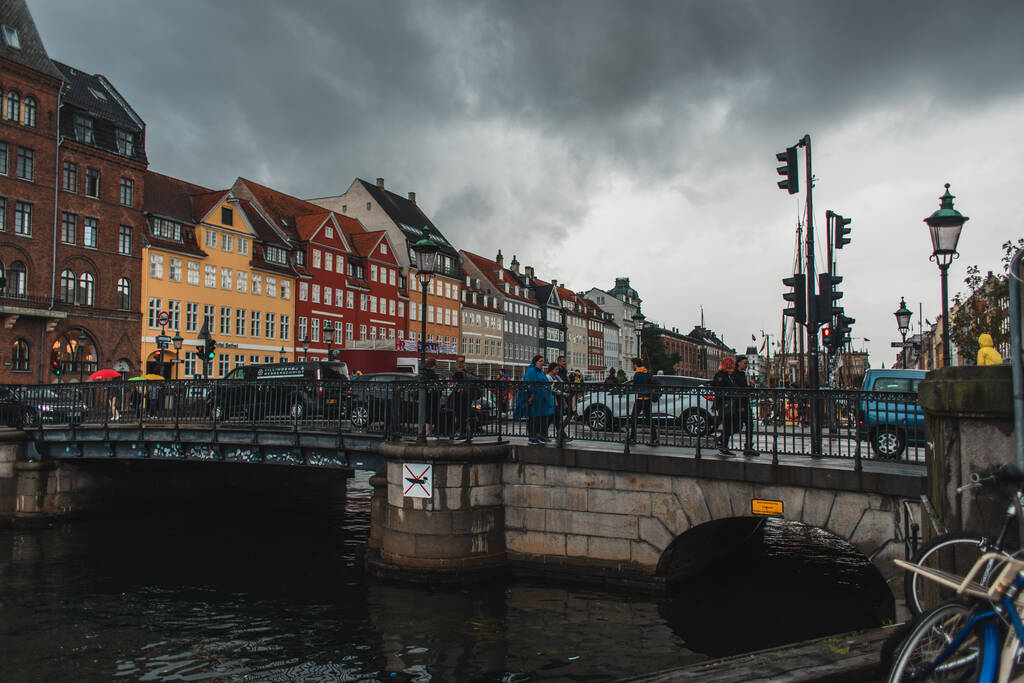 COPENHAGEN, DENMARK - 30 Nisan 2020: Nyhavn kentsel caddesi ve arka planda bulutlu gökyüzü ile kanal yakınlarındaki köprüde yürüyen insanlar   - Fotoğraf, Görsel