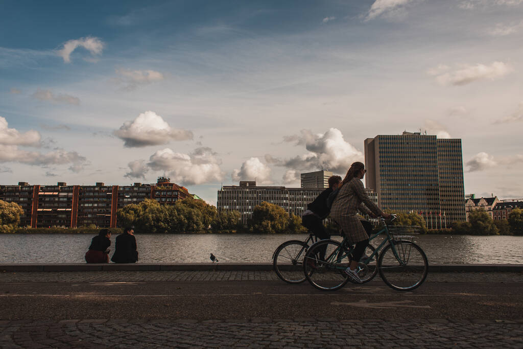 COPENAGEN, DANIMARCA - 30 APRILE 2020: Persone che pedalano in bicicletta sul lungomare vicino al canale con edifici e cielo nuvoloso sullo sfondo  - Foto, immagini