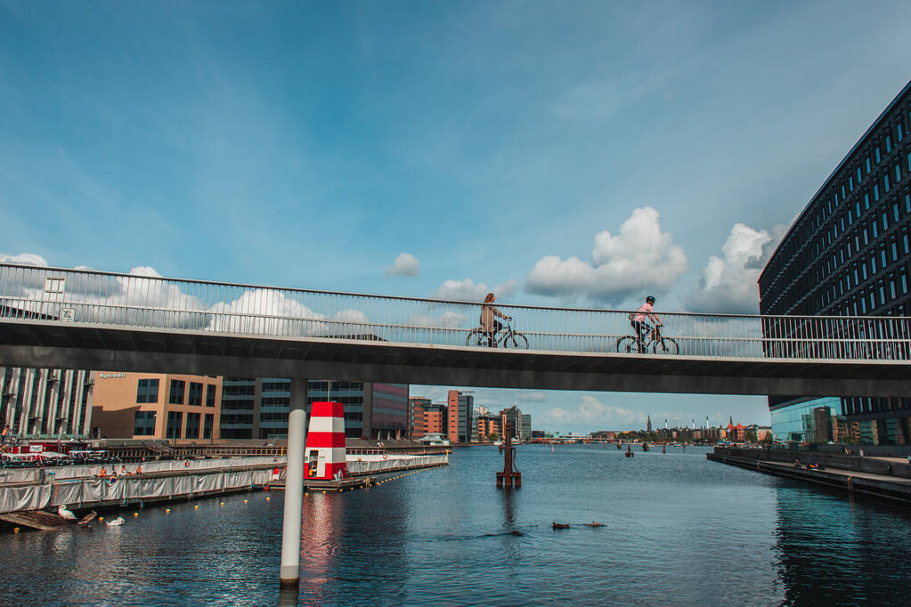 COPENHAGEN, DENMARK - APRIL 30, 2020: Люди їздять на велосипедах по мосту над річкою з будівлями та хмарним небом на задньому плані.   - Фото, зображення