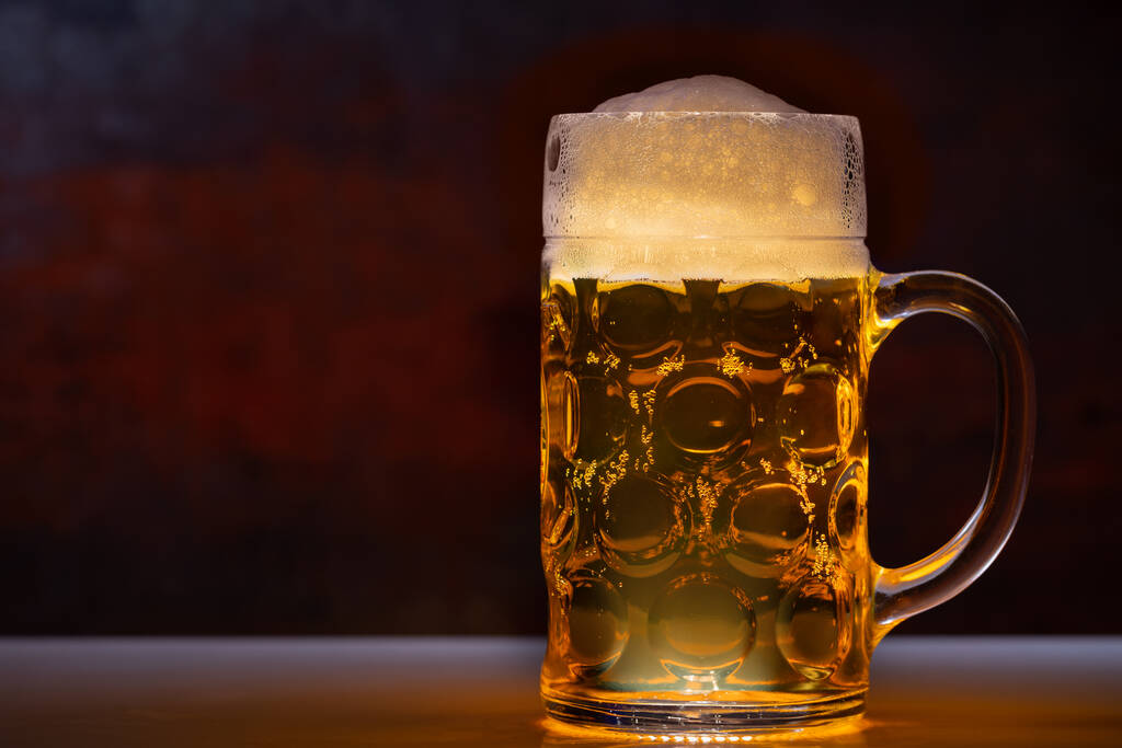 Tankard cheio de cerveja com uma cabeça espumosa em uma visão lateral close-up em um balcão com espaço de cópia para o lado iluminado com luz
 - Foto, Imagem