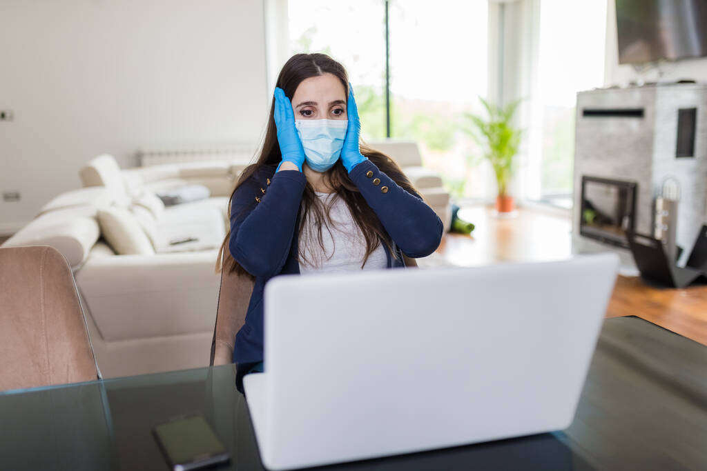 Gestresste vrouw met gezichtsmasker en beschermende handschoenen tijdens het werken op laptop thuis. Covid-19 en zakelijke problemen concept. - Foto, afbeelding