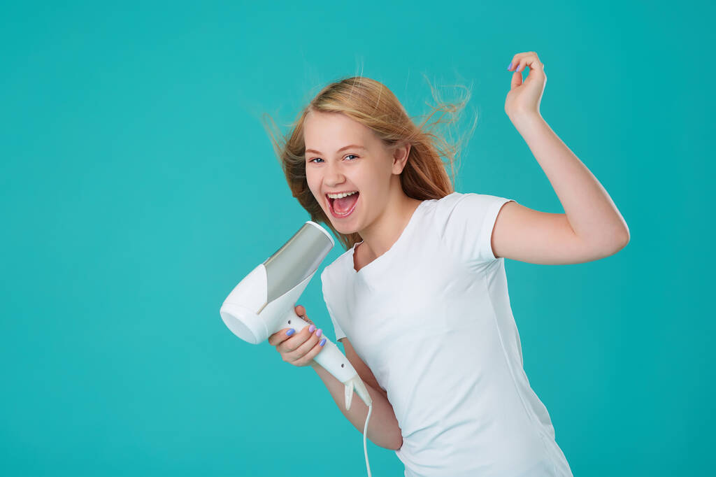 Una felice ragazza allegra si asciuga i capelli con un asciugacapelli e canta usando l'asciugacapelli come microfono. Sfondo blu menta, Ritratto Studio
. - Foto, immagini