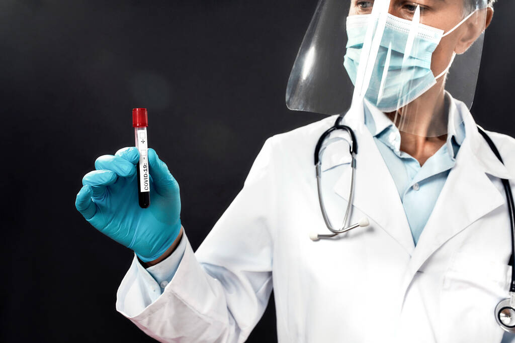Reifer männlicher Arzt mit medizinischer Maske und Gesichtsschutz, der das Reagenzglas mit Blut für den COVID-19-Test hält. Schwerpunkt im Reagenzglas. Labortests auf Coronavirus - Foto, Bild
