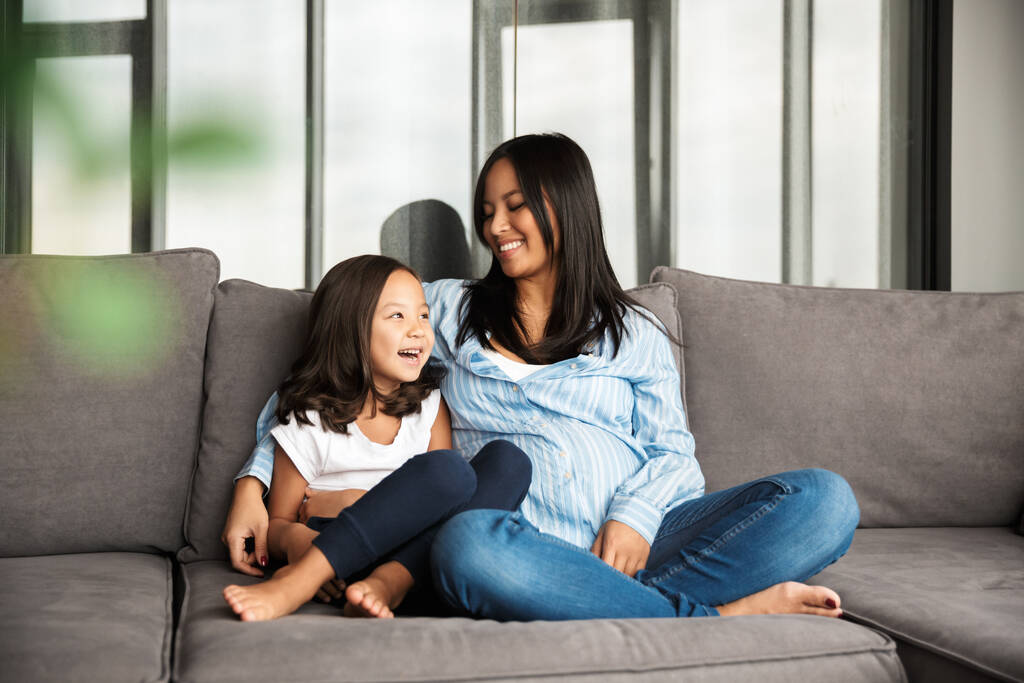 Фото щасливої вагітної азіатки з її маленькою дочкою обіймається і сміється, сидячи на дивані вдома
 - Фото, зображення