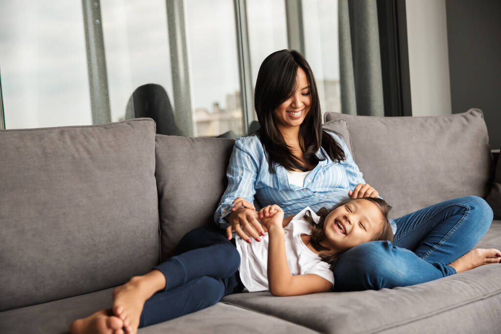 Foto de feliz mulher asiática grávida com sua filhinha se divertindo e rindo enquanto descansa no sofá em casa
 - Foto, Imagem