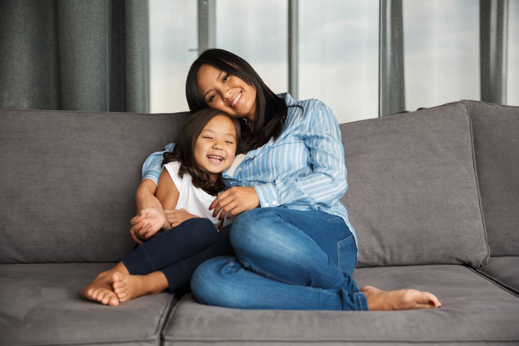Photo of pleased pregnant asian woman with her little daughter hugging and smiling while sitting on couch at home - Photo, Image