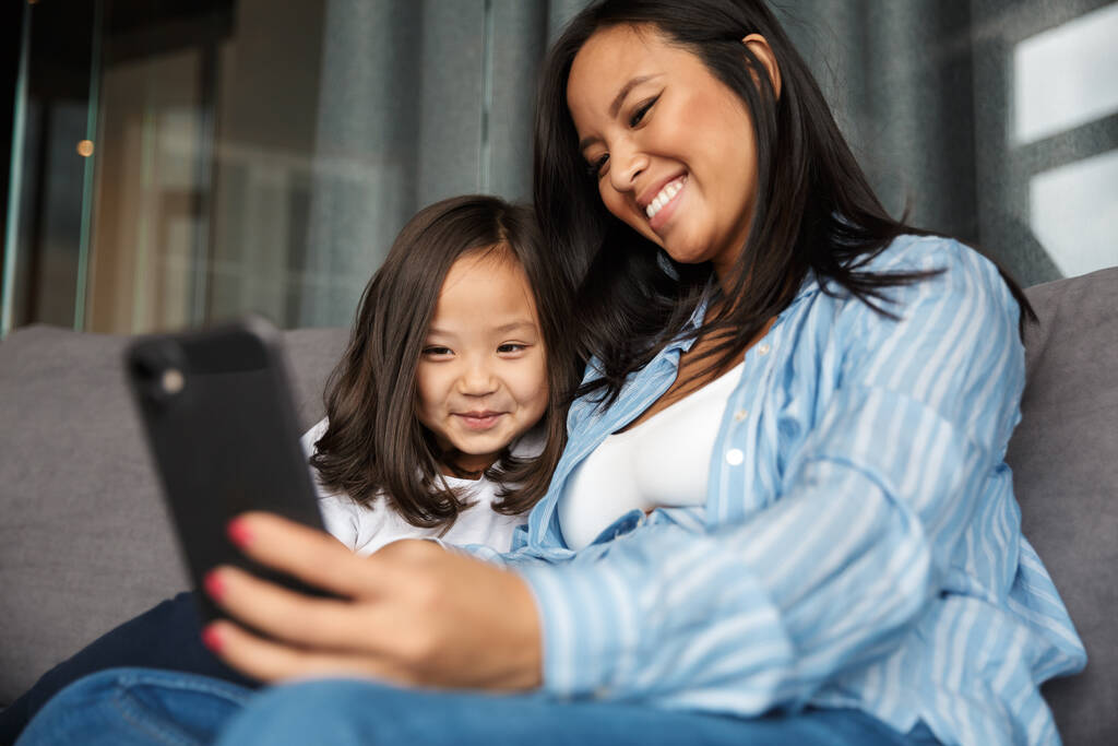 Foto van vrolijke zwangere aziatische vrouw met haar kleine dochter glimlachen en het gebruik van mobiele telefoon terwijl zitten op de bank thuis - Foto, afbeelding