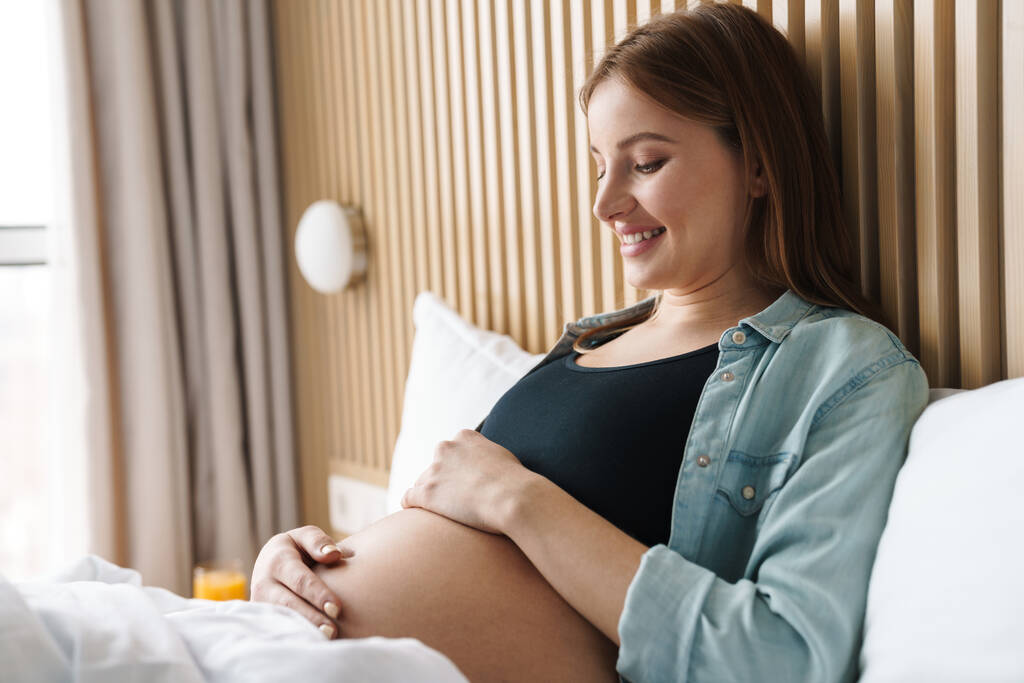 Foto von glücklich schwangeren Frau mit roten Haaren lächelnd und streichelt ihren Bauch, während sie auf dem Bett in sonnigem Zimmer liegt - Foto, Bild
