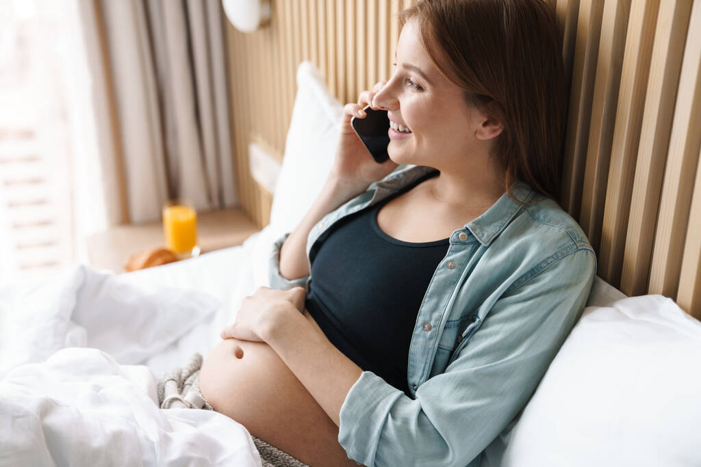 Foto van gelukkige zwangere vrouw met rood haar glimlachen en praten op mobiele telefoon terwijl ze op bed in de zonnige kamer - Foto, afbeelding