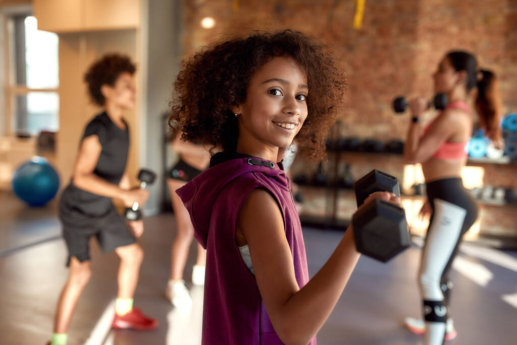 Afroamerykanka uśmiechnięta do kamery podczas treningu z hantlami na siłowni wraz z trenerką i innymi dziećmi. Sport, zdrowy styl życia, wychowanie fizyczne - Zdjęcie, obraz