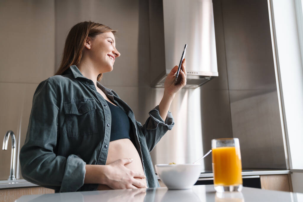 Фотографія радісної вагітної жінки з рудим волоссям за допомогою мобільного телефону під час сніданку на сучасній кухні
 - Фото, зображення