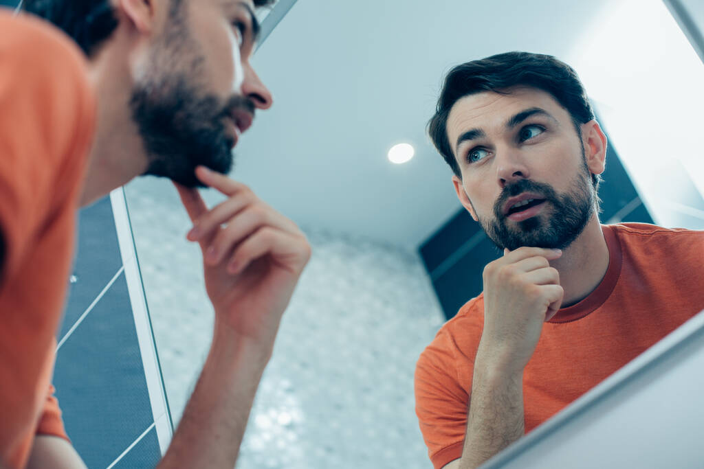 Rauhallinen utelias nuori mies katselee tarkkaavaisesti peilikuvaansa ja koskettaa partaansa seistessään kylpyhuoneessa - Valokuva, kuva