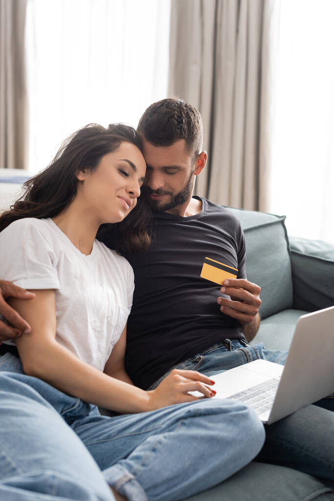 hombre guapo sosteniendo la tarjeta de crédito y abrazando novia feliz cerca del ordenador portátil
  - Foto, imagen