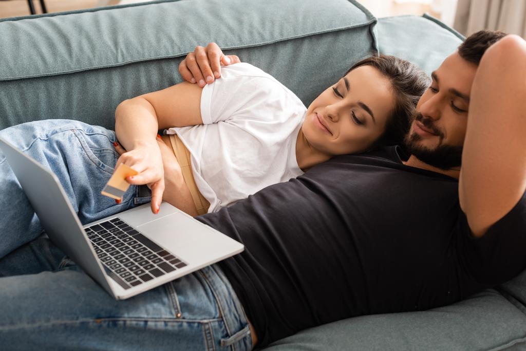 attraktive Frau liegt mit hübschem Freund auf Sofa und hält Kreditkarte in der Nähe von Laptop  - Foto, Bild
