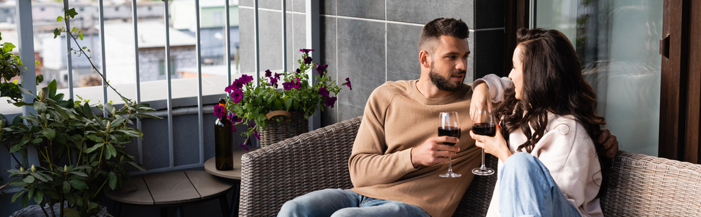 panoramisches Konzept eines gut aussehenden Mannes und einer attraktiven Frau, die auf einem Sofa im Freien sitzen und Weingläser halten, während sie einander anschauen - Foto, Bild