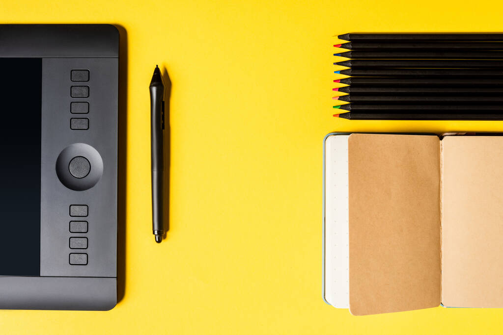 Top view of graphics tablet and stylus near open notebook and color pencils on yellow surface - Photo, Image