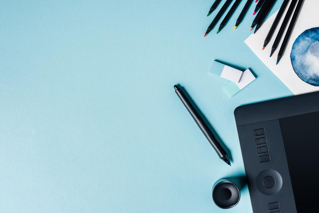 Верхний вид графического планшета со стилусом, цветными карандашами и акварелью на синем фоне
 - Фото, изображение