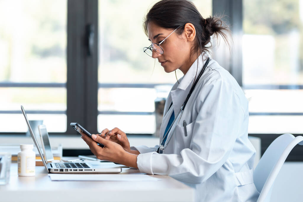 Снимок серьезной женщины-врача с помощью смартфона во время работы с ноутбуком в консультации
. - Фото, изображение