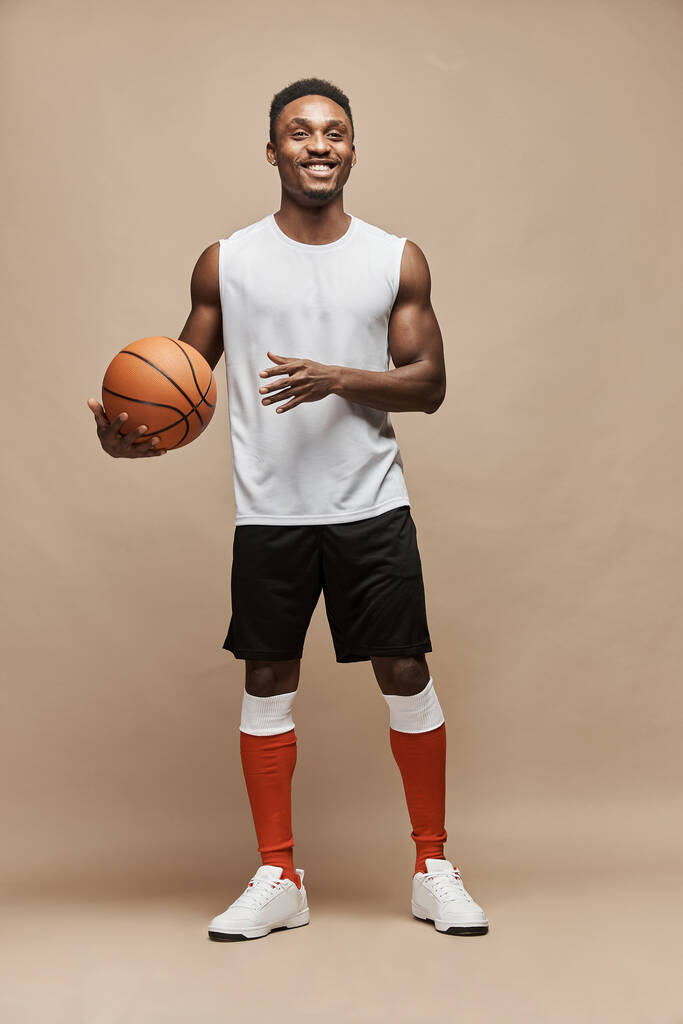 πλήρης φωτογραφία ενός μαύρου αθλητικού μπασκετμπολίστα στο στούντιο σε μπεζ φόντο φορώντας λευκό μπλουζάκι, μαύρο σορτς, κόκκινες μακριές κάλτσες και λευκά sneakers, κρατάει την μπάλα στο χέρι του και χαμογελάει - Φωτογραφία, εικόνα