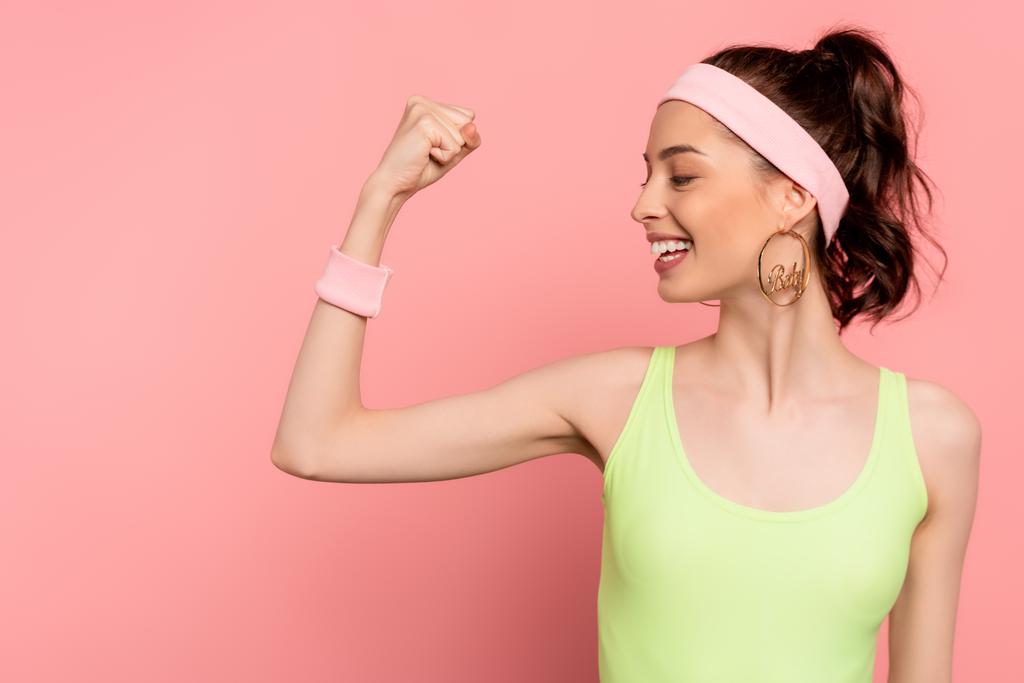 Χαρούμενη αθλήτρια με σφιχτή γροθιά κοιτάζοντας τους μυς στο ροζ  - Φωτογραφία, εικόνα