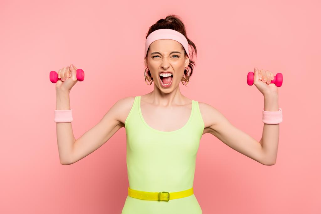 emotionele sportvrouw kijken naar camera en oefenen met halters op roze  - Foto, afbeelding