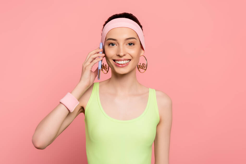 Fröhliche Frau mit Stirnband spricht auf Smartphone isoliert auf rosa  - Foto, Bild