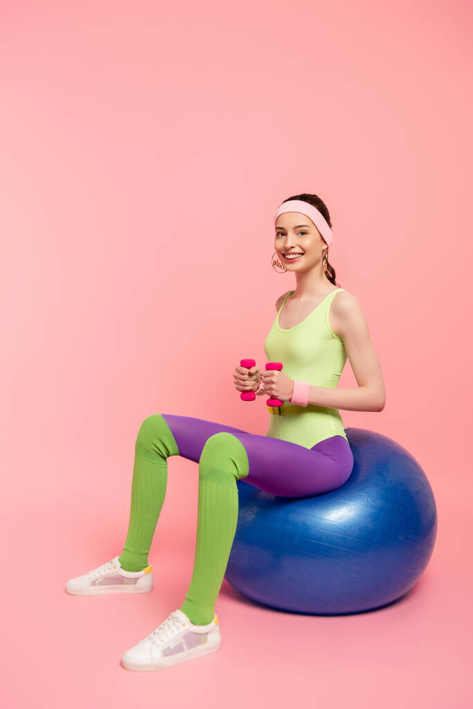 gelukkige sportvrouw zittend op fitness bal en oefenen met halters op roze  - Foto, afbeelding