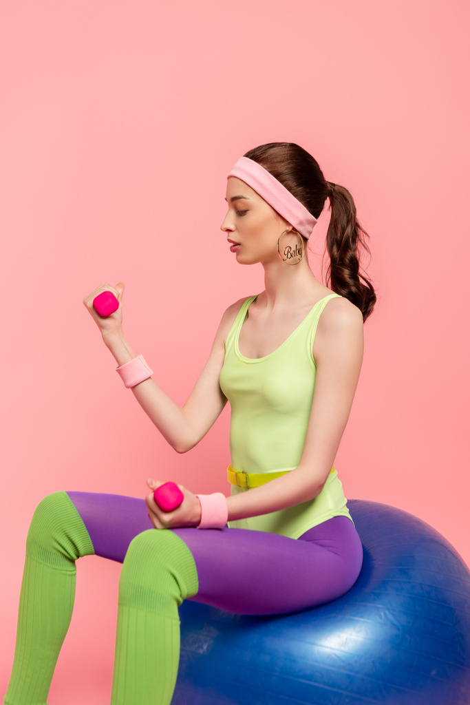 молодая спортсменка сидит на фитнес-мяч и упражнения с гантели изолированы на розовый
  - Фото, изображение
