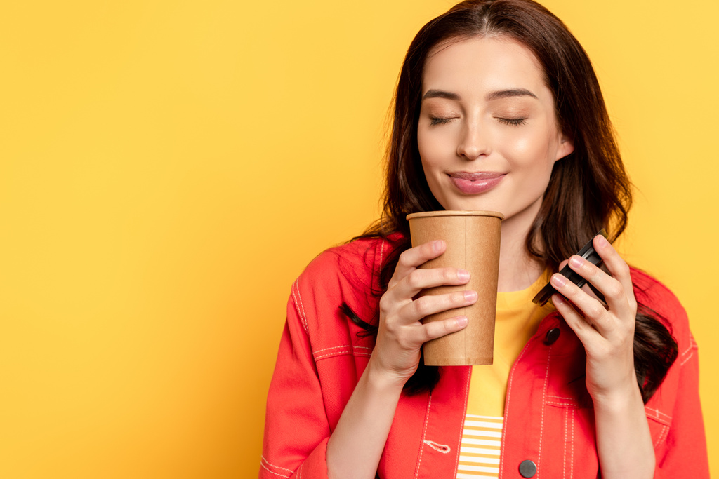 明るい若い女性が紙コップを持ってコーヒーの匂いを嗅いで黄色に隔離され - 写真・画像