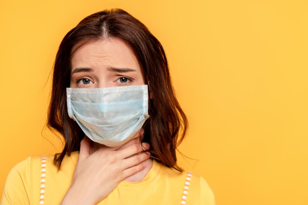 zieke jonge vrouw in medisch masker aanraken hals terwijl het hebben van zere keel geïsoleerd op geel - Foto, afbeelding