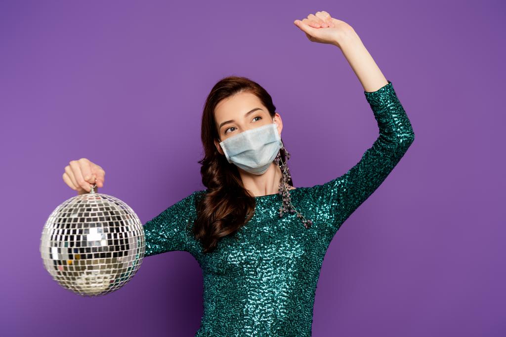 vrouw in jurk en medisch masker houden disco bal op paars - Foto, afbeelding
