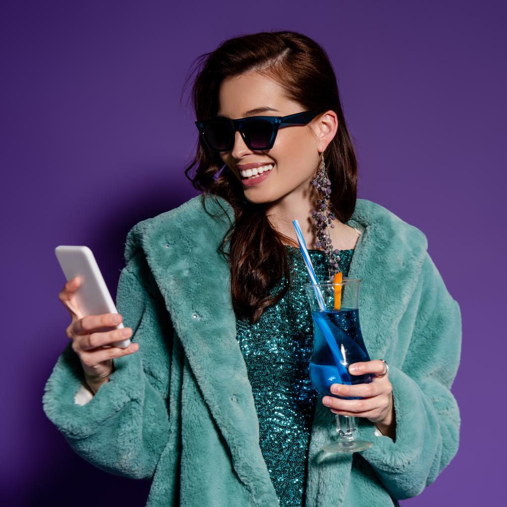 fröhliche Frau mit Sonnenbrille hält Glas mit Cocktail und Smartphone auf lila  - Foto, Bild