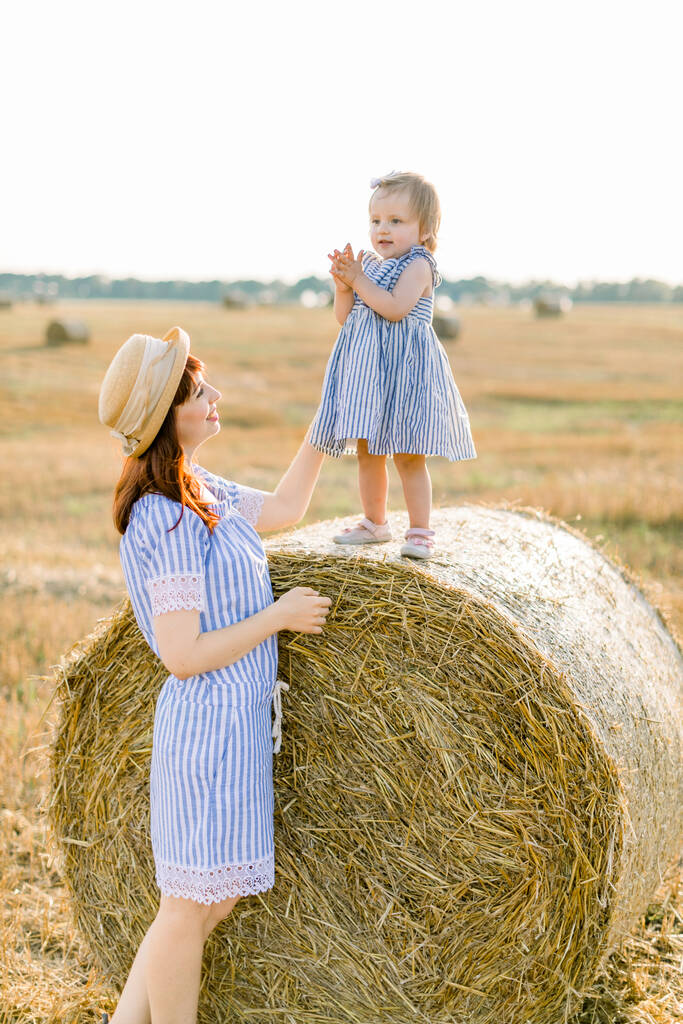 Happy mother and little daughter, wearing similar striped dresses, having fun in summer field with golden hay bales. Child on a haystack. Countryside, happy childhood - Valokuva, kuva