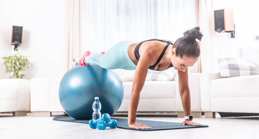 Jong atletisch meisje houdt een plank positie op haar woonkamer, met benen op de top van de fitness bal met een fles water, twee halters en een mat in de kamer. - Foto, afbeelding
