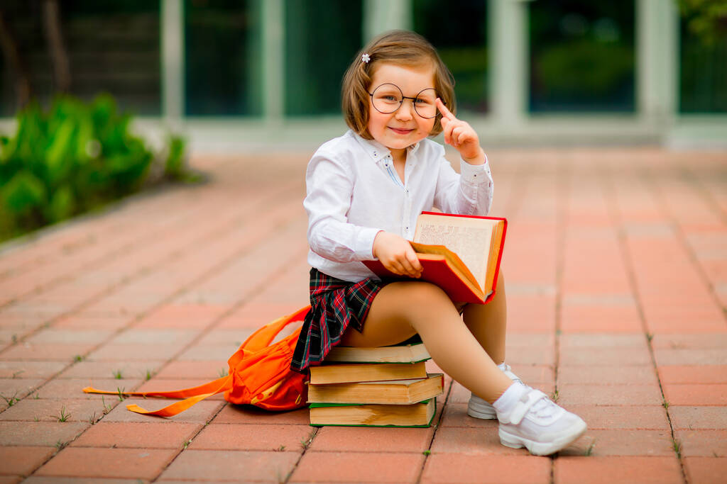 una bambina in uniforme scolastica e occhiali, seduta su una pila di libri vicino alla scuola
 - Foto, immagini